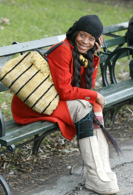 Erykah Badu sitting on a park bench, red coat, black hat, and pigtail wig
