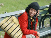 Erykah Badu sitting on a park bench, red coat, black hat, and pigtail wig